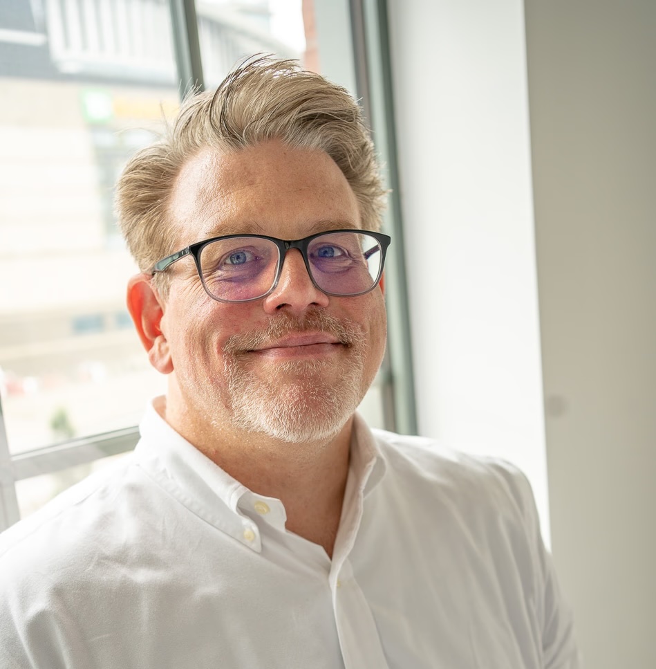 Don, a man with short grey hair, a trimmed grey beard and dark-framed glasses, smiling by a bright window